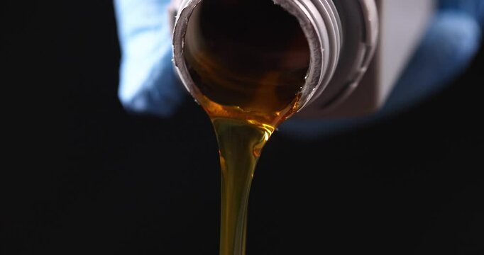 Person pours stream of golden-brown motor oil from container into engine. Viscous fluid catches light as flows indicating texture and consistency