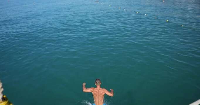 Man in blue swim trunks runs and jumps energetically into sea from pier. Arms and legs spread wide in expression of excitement on sunny day