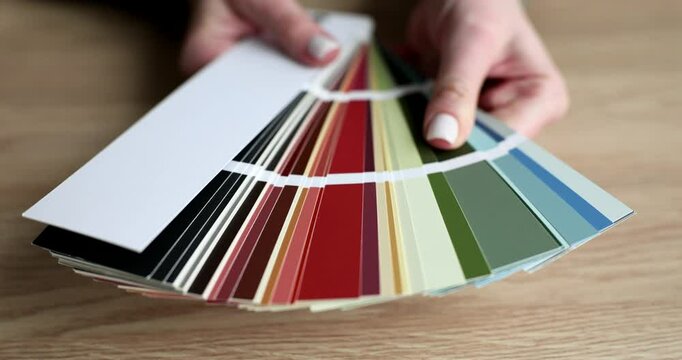 Woman holds fan deck of color swatches over wooden table. Swatches display wide range of colors suggesting selection process for interior decoration