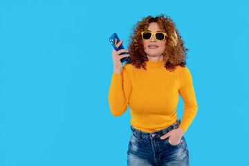 Young woman with curly hair wearing sunglasses and a bright yellow sweater holds a smartphone against a vibrant blue background while posing confidently