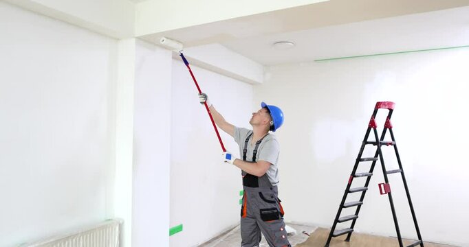 Craftsman uses roller to paint room ceiling with white paint. Construction worker applies fresh coat of paint with smooth and steady strokes
