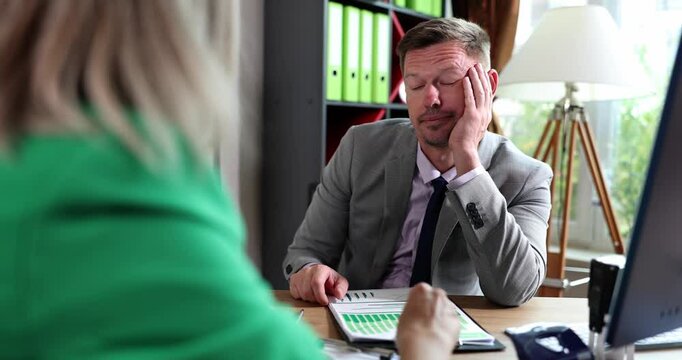 Man in suit sits at desk opposite woman discussing project and reviewing papers. Office workers look bored and disinterested in workplace meeting