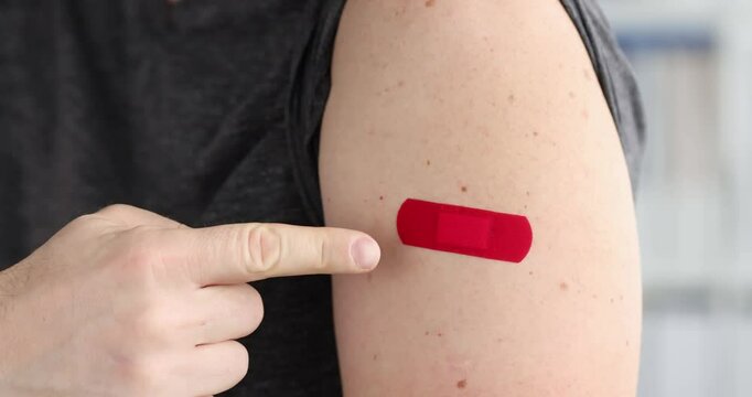 Man rolls up t-shirt sleeve showing patch on arm in light room. Patient demonstrates vaccination bandage on skin in healthcare facility