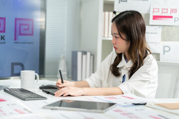 Smiling Young asian Designer Working design logo brand on Tablet in Bright Modern Office Space,Surrounded by branding designs.Asian Designer Woman Taking Business Call at Creative Office Desk