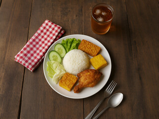 A traditional Asian meal featuring rice, fried chicken, vegetables, tempeh, and a refreshing iced tea, served on a rustic wooden table with a gingham cloth napkin and silver cutlery.