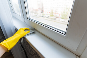 A person who is wearing bright yellow gloves is diligently cleaning the window sill to ensure a spotless and tidy appearance in their home environment while promoting cleanliness