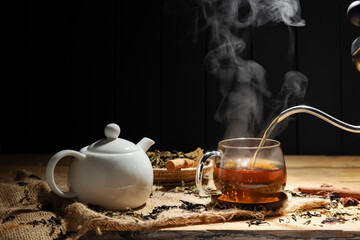 Pour hot tea into cups, herbal tea glasses are arranged on a rustic wooden table.