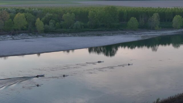 Rowing boats leaves rippling trails over the mirror like Po river, flanked by a sandbar and dense riverside forest, as reflections soften the early light, amplifying tranquil atmosphere, slow motion