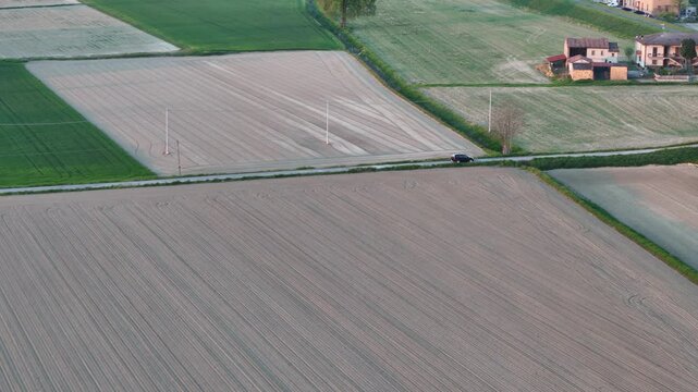 Drone glides over patterned farmland near Olza di Castelvetro Piacentino, PC, as a black suv moves along a rural road bordered by tilled soil, green pastures, and scattered country homes, slow motion