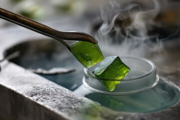 Green shimmering cubes held by metal tongs over frosted glass container with visible vapor and condensation