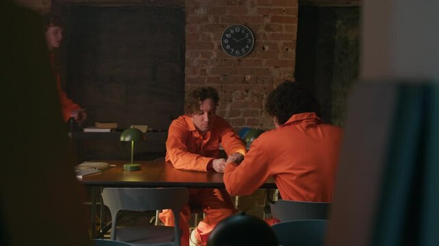 Male prisoner showing awl to cellmate, slipping him it and pretending to read sitting at table opposite each other when another guy walking past them with book trolley in library