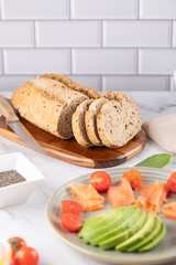 Sliced whole grain bread on wooden board next to a plate with smoked salmon, avocado and cherry tomatoes. Bright kitchen background. Healthy breakfast concept