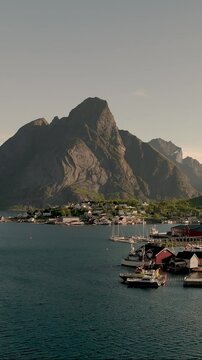 Breathtaking coastal landscape showcasing majestic rocky mountains overlooking a tranquil harbor in Norway. Sailboats are gently anchored, Reine, Lofote,n Norway