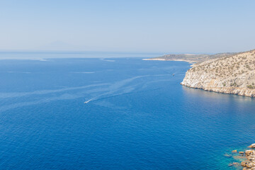 Expansive view of the deep blue sea contrasting with a rugged, rocky coastline. A small boat's wake...