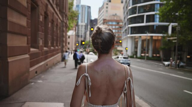 A woman in a light, backless dress walks along a city street in Sydney, surrounded by a mix of historic sandstone buildings and modern glass skyscrapers.