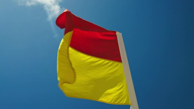 A red and yellow flag waves in the wind against a blue sky, marking designated swimming zones on Australian beaches. Essential coastal safety symbol protecting beachgoers in surf conditions.
