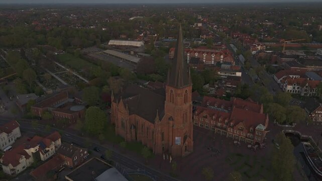 Orbiting drone shot of the red-brick St. Antonius Church in Papenburg, Germany, during golden hour. Surrounded by rooftops, trees and evening light. Historic architecture in a peaceful townscape.