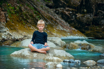 Blond boy sits cross-legged in lotus pose on a rock in the middle of a fast mountain river. Peaceful scene of childhood, nature, meditation and freedom in the wild.