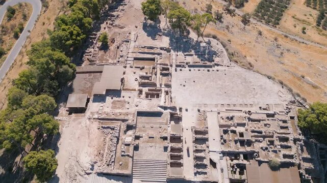 Aerial drone footage reveals the ancient Minoan ruins of Phaistos in Crete. Historic site with stone structures, mountain views, and cultural significance