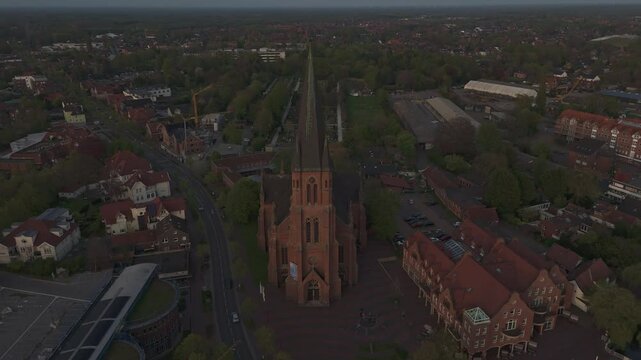 Orbiting drone shot of the red-brick St. Antonius Church in Papenburg, Germany, during golden hour. Surrounded by rooftops, trees and evening light. Historic architecture in a peaceful townscape.