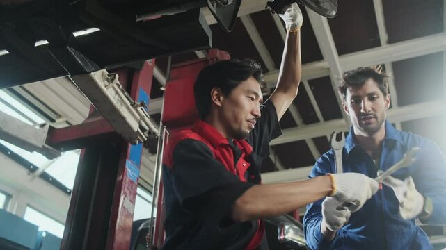 Two male mechanics cooperate, Latin and Asian workers fixing automotive wheels and maintaining car suspensions in garage station, and technician engineering service jobs in vehicle transport industry.