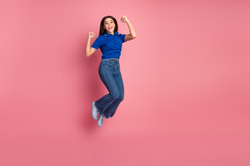 Happy young woman jumping excitedly in a blue shirt and jeans against a pink background, expressing...