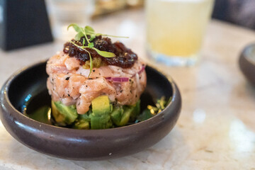 Delicious seafood salmon tartare sits atop creamy avocado in a modern restaurant environment. The dish is garnished with a rich sauce, showcasing