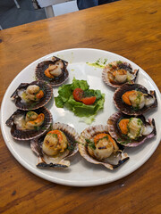 A top-down shot of grilled scallops in their shells, artfully arranged on a white plate with fresh garnishes.