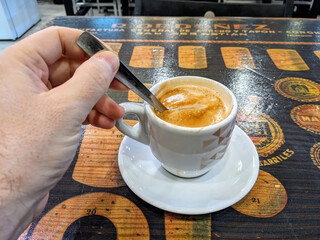 A hand stirring a hot espresso coffee drink in a white cup, sitting on a rustic, vintage table.