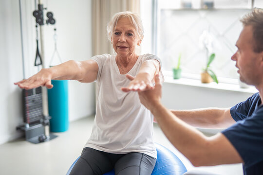 Senior woman doing physical therapy exercises with physiotherapist on swiss ball. Elderly woman exercising with personal trainer using fitness ball in rehabilitation center