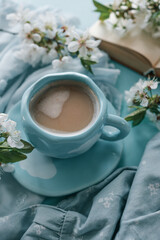 A cup of coffee with foam in a blue cup with white cloud design, surrounded by white spring blossoms, a soft blue fabric, and an open book, evoking a serene and cozy atmosphere