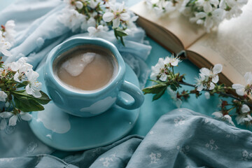 A cup of coffee with foam in a blue cup with white cloud design, surrounded by white spring blossoms, a soft blue fabric, and an open book, evoking a serene and cozy atmosphere