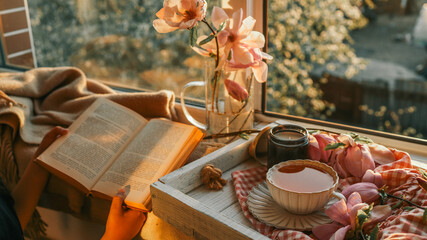 Person reading a book by a cozy window at sunset, with magnolia flowers, a black candle, and a cup of tea on a wooden tray. Warm golden light creates a peaceful, serene mood