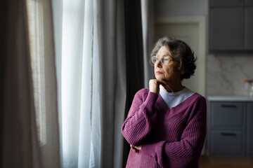 Depressed upset elderly woman gazing out of window with thoughtful expression, feeling lonely and...