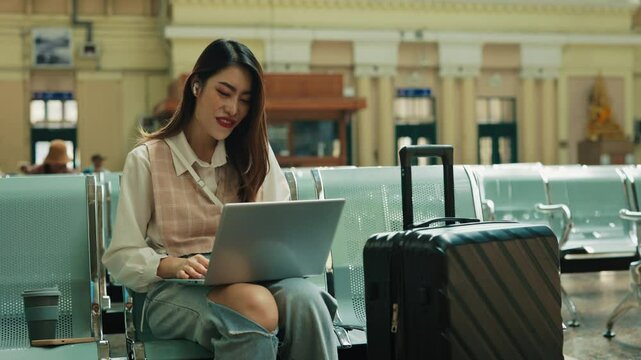 Portrait of excited Asian lesbian talking via video call in waiting hall of train station. Friendly trans woman waves hand to laptop camera sitting on chair. Journey on holiday