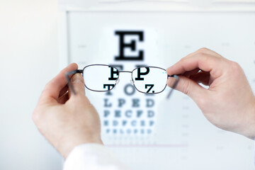Man holding glasses against eye chart on light wall background, closeup shot. Ophthalmologist...
