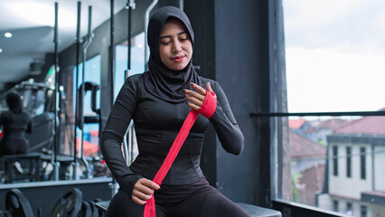 A Southeast Asian Muslim woman in a black sports hijab wraps her hand with red boxing tape while sitting in a gym. The moment shows strength, preparation, and confidence in a modern wellness