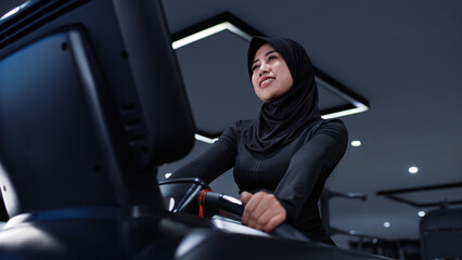 A smiling Southeast Asian Muslim woman in a black sports hijab works out on a treadmill in a modern gym. The photo reflects positivity, health, and the joy of movement in an inclusive fitness  © Sergei