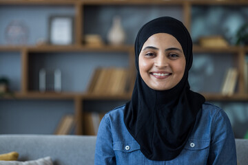 Close-up portrait of a young Muslim woman in a black hijab sitting on a sofa at home and smiling at the camera