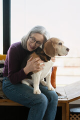 Senior woman embracing beagle dog sitting near window at home