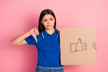 Young female in casual blue top showing mixed approval and thumb-down gesture holding sign on pink...