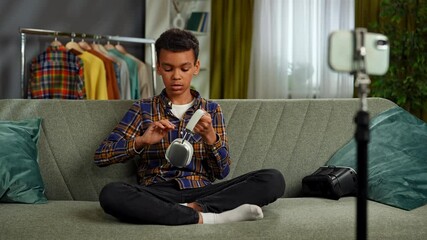 Young african american child boy spending time at home, kid blogger sits on sofa records video on smartphone, talking at camera showing headphones and gives review.