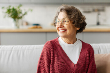 Happy grey-haired senior woman sitting on couch at home and looking aside at free space with smile,...