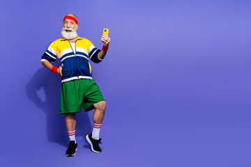 Elderly man in retro sportswear posing with a smartphone against a purple background showing active and healthy lifestyle