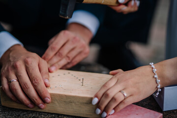 Obraz premium Close-up of bride and groom hammering nails into a wooden block during a symbolic wedding ritual, showcasing hands, rings, and shared effort..