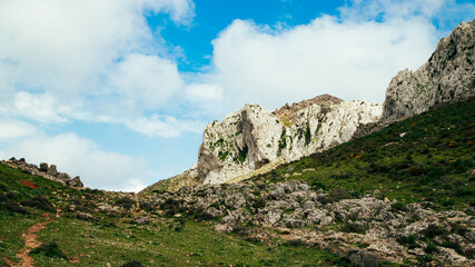 Mountainous Moroccan landscape, bright white rocks, green hills, blue sky scattered with clouds, discreet hiking trail winding through the wild nature.