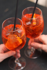 Two glasses with Aperol Spritz in the hands of girls. Cold refreshing alcoholic cocktail Aperol Spritz. Cocktails close-up with selective focus. Alcoholic drink with a straw. Traditional Italian drink