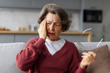 Depressed European senior woman suffering from headache touching head, sitting on couch in living room at home, having mental health issues and depression