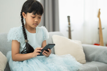 Young girl with braid sitting on a couch, focused on mobile device, wearing light blue dress in cozy living room with natural light and musical instruments