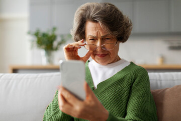 Elderly European woman squinting eyes trying read message on cellphone screen, sitting on couch at...